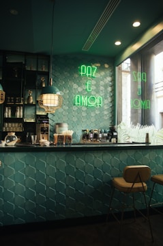A cozy caf&eacute; interior with teal geometric wall tiles and a countertop equipped with coffee machines and various supplies. The space is lit with stylish pendant lights and has a neon sign in green reading 'PAZ E AMOR' on the wall. Two warm-colored stools are positioned at the bar, creating a welcoming atmosphere.