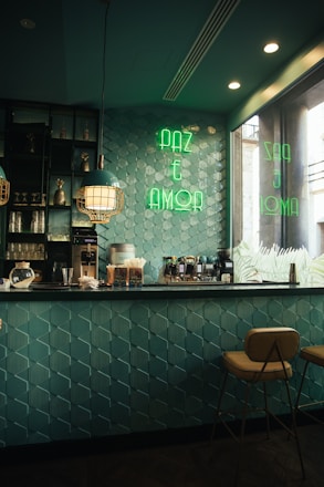 A cozy caf&eacute; interior with teal geometric wall tiles and a countertop equipped with coffee machines and various supplies. The space is lit with stylish pendant lights and has a neon sign in green reading 'PAZ E AMOR' on the wall. Two warm-colored stools are positioned at the bar, creating a welcoming atmosphere.