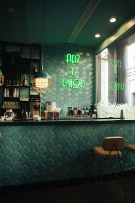 A cozy café interior with teal geometric wall tiles and a countertop equipped with coffee machines and various supplies. The space is lit with stylish pendant lights and has a neon sign in green reading 'PAZ E AMOR' on the wall. Two warm-colored stools are positioned at the bar, creating a welcoming atmosphere.