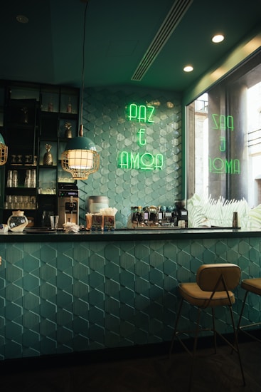 A cozy caf&eacute; interior with teal geometric wall tiles and a countertop equipped with coffee machines and various supplies. The space is lit with stylish pendant lights and has a neon sign in green reading 'PAZ E AMOR' on the wall. Two warm-colored stools are positioned at the bar, creating a welcoming atmosphere.
