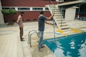 Two people are beside a swimming pool, one of whom is using a cleaning tool with a long handle to clean the pool. The environment includes a diving board, tiled floor, and a red brick wall. A garden area with a tree is visible in the background.