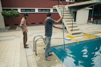 Two people are beside a swimming pool, one of whom is using a cleaning tool with a long handle to clean the pool. The environment includes a diving board, tiled floor, and a red brick wall. A garden area with a tree is visible in the background.