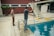 Two people are beside a swimming pool, one of whom is using a cleaning tool with a long handle to clean the pool. The environment includes a diving board, tiled floor, and a red brick wall. A garden area with a tree is visible in the background.