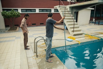 Two people are beside a swimming pool, one of whom is using a cleaning tool with a long handle to clean the pool. The environment includes a diving board, tiled floor, and a red brick wall. A garden area with a tree is visible in the background.