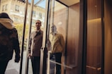 A well-dressed hotel doorman is holding open a glass door for a person entering the building. The scene is bright with sunlight filtering in, and parts of a building facade are visible outside.