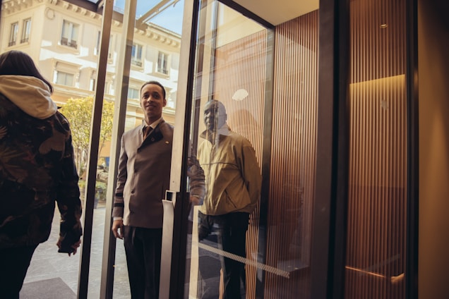 A well-dressed hotel doorman is holding open a glass door for a person entering the building. The scene is bright with sunlight filtering in, and parts of a building facade are visible outside.