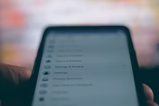 A close-up of a smartphone screen displaying a settings and privacy menu. The text appears slightly blurred, with options like settings, privacy shortcuts, and terms and policies visible. The background is out of focus, creating a contrast with the darker foreground.