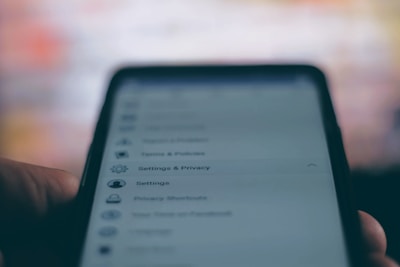 A close-up of a smartphone screen displaying a settings and privacy menu. The text appears slightly blurred, with options like settings, privacy shortcuts, and terms and policies visible. The background is out of focus, creating a contrast with the darker foreground.