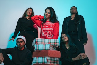 A group photo features five individuals with a casual and festive atmosphere. The central figure is wearing a red holiday sweater and is surrounded by colorful, wrapped gift boxes. One person is wearing a Santa hat, and the lighting casts a green and red hue over the scene.