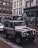 A rugged white off-road vehicle is parked on a cobblestone street in an urban setting. The vehicle has large tires and appears to be a Land Rover Defender. The street is lined with modern and old-style buildings, featuring large windows and various storefronts. Pedestrians are seen in the background, and other cars are parked along the street. The weather seems overcast, and the scene has a somewhat muted color palette.