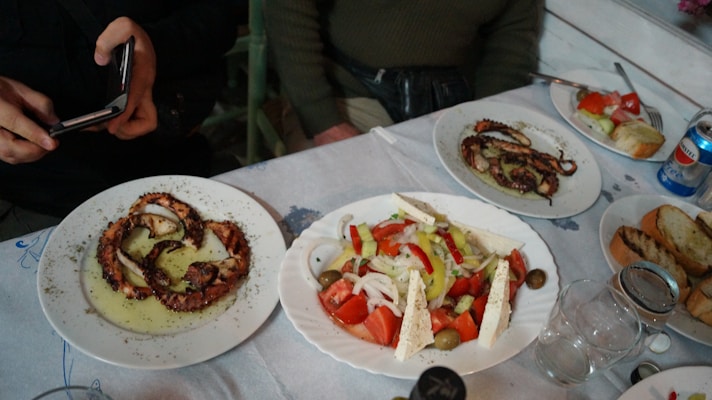 A table set with plates of food, featuring a dish of grilled octopus in olive oil, a Greek salad with tomatoes, cucumbers, onions, olives, and feta cheese, and slices of toasted bread. A person holding a smartphone is capturing the food, and there are glasses and a can of soda on the table.