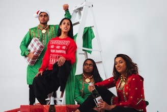 A group of four people dressed in festive attire, including colorful sweaters and accessories, posing with a wrapped gift and sitting near a white ladder. They appear to be celebrating, possibly during a holiday season.