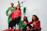 A group of four people dressed in festive attire, including colorful sweaters and accessories, posing with a wrapped gift and sitting near a white ladder. They appear to be celebrating, possibly during a holiday season.