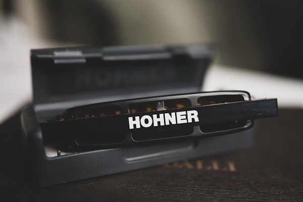 A close-up view of a harmonica positioned on top of its open case. The harmonica has the brand name 'Hohner' written prominently on it and is placed against a blurred background.