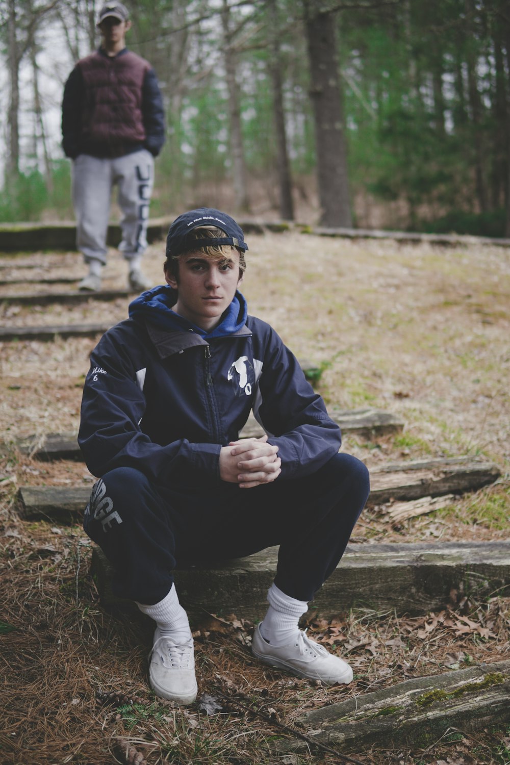 man sitting on gray log near another man standing surrounded with tall and green trees