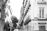 A narrow street scene featuring tall buildings with wrought iron balconies. A large tree is on the left, and a street sign reads 'Calle de San Andrés'. A shop sign advertising 'gelato y orxata' is visible, and there is a parked car along the street.
