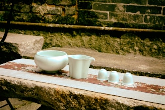 Several pieces of white ceramic, including a bowl, a jug, and three small cups, are arranged on a rustic stone surface. The background consists of a weathered brick wall and patches of greenery, giving the scene an antique and serene ambiance.