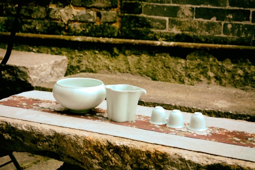 Several pieces of white ceramic, including a bowl, a jug, and three small cups, are arranged on a rustic stone surface. The background consists of a weathered brick wall and patches of greenery, giving the scene an antique and serene ambiance.