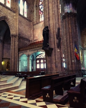 A grand interior of a cathedral featuring high vaulted ceilings with intricate stonework. Stained glass windows cast colorful light into the space. Wooden pews arranged in rows lead towards a raised platform. A statue draped in fabric is attached to a column next to a flag.