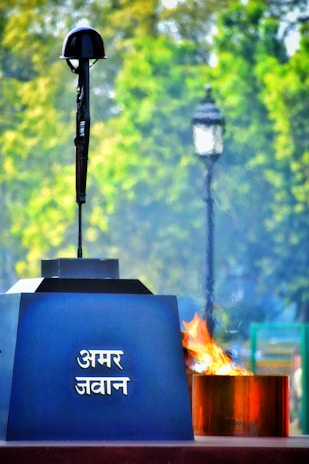 A memorial structure includes a rifle standing vertically with a helmet on top and an eternal flame beside it. The background displays greenery, and a lamp post is visible. The inscription on the structure is in a script that appears to be Hindi.