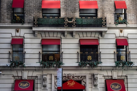 A classic urban building facade features several windows with red awnings. Ornate black iron railings adorn the windows, with decorative golden emblems. Greenery and red flowers line the railings, adding a touch of natural beauty. The building also displays signage for 'Tender' in circular plaques.