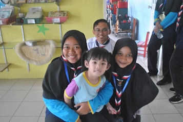 A group of five people, including a young child and four others, are gathered indoors. The individuals are smiling and appear to be enjoying each other's company. They are wearing scout uniforms, indicating some association with a scouting or guiding organization. Shelves with different items, such as books and a plush item, are visible in the background.
