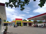 A gas station featuring a modern design with a bold color scheme of red, green, and white. The forecourt area includes petrol pumps, a parked car, and a small building with signage. The sky is bright with scattered clouds, and some green trees are visible around the premises. The ground is paved with gray tiles and a few potted plants are placed near the entrance.