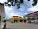 A gas station featuring a modern design with a bold color scheme of red, green, and white. The forecourt area includes petrol pumps, a parked car, and a small building with signage. The sky is bright with scattered clouds, and some green trees are visible around the premises. The ground is paved with gray tiles and a few potted plants are placed near the entrance.