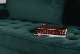 A vintage book with a red-brown cover and a worn spine rests on top of a notebook with a spiral binding. These items are placed on a plush, dark green tufted couch.