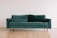A green velvet sofa with wooden legs positioned in a minimalist room featuring light wooden flooring and a plain white wall in the background.