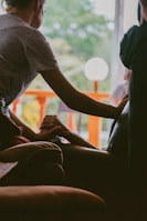 A person gently holds the hand of another seated individual, conveying a sense of comfort and support. The interaction takes place indoors, with a bright window in the background revealing green foliage and an orange railing. The lighting is soft and creates a warm atmosphere.