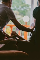 A person gently holds the hand of another seated individual, conveying a sense of comfort and support. The interaction takes place indoors, with a bright window in the background revealing green foliage and an orange railing. The lighting is soft and creates a warm atmosphere.