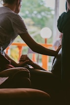 A person gently holds the hand of another seated individual, conveying a sense of comfort and support. The interaction takes place indoors, with a bright window in the background revealing green foliage and an orange railing. The lighting is soft and creates a warm atmosphere.