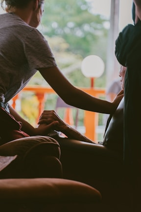 A person gently holds the hand of another seated individual, conveying a sense of comfort and support. The interaction takes place indoors, with a bright window in the background revealing green foliage and an orange railing. The lighting is soft and creates a warm atmosphere.
