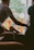 A person gently holds the hand of another seated individual, conveying a sense of comfort and support. The interaction takes place indoors, with a bright window in the background revealing green foliage and an orange railing. The lighting is soft and creates a warm atmosphere.