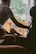 A person gently holds the hand of another seated individual, conveying a sense of comfort and support. The interaction takes place indoors, with a bright window in the background revealing green foliage and an orange railing. The lighting is soft and creates a warm atmosphere.