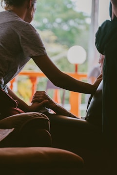 A person gently holds the hand of another seated individual, conveying a sense of comfort and support. The interaction takes place indoors, with a bright window in the background revealing green foliage and an orange railing. The lighting is soft and creates a warm atmosphere.