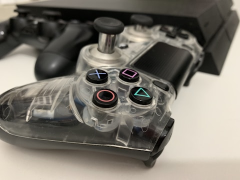 A clear gaming controller is resting on a white surface, showcasing its transparent casing with visible internal components. Nearby, there is a black console, partially blurred, along with a second black controller slightly out of focus.