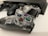 A clear gaming controller is resting on a white surface, showcasing its transparent casing with visible internal components. Nearby, there is a black console, partially blurred, along with a second black controller slightly out of focus.