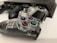 A clear gaming controller is resting on a white surface, showcasing its transparent casing with visible internal components. Nearby, there is a black console, partially blurred, along with a second black controller slightly out of focus.