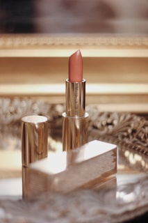 A lipstick with a beige-pink shade is standing upright on a reflective, ornate surface. The packaging is gold with the brand name 'BECCA' visible on it. The background shows a blurred golden frame, contributing to a luxurious setting.