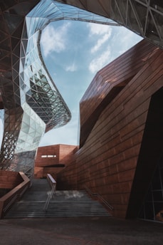 Futuristic architectural structure featuring a combination of glass and rust-colored metal with dynamic, angular shapes and geometric patterns. The design includes a large staircase leading upwards, with the sky visible through the interwoven structures.