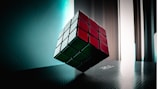 A Rubik's Cube is suspended in mid-air, captured in a softly lit room with light coming from tall windows. The cube displays red, white, and green stickers, and is positioned at an angle on a textured surface with a reflective sheen.