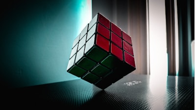 A Rubik's Cube is suspended in mid-air, captured in a softly lit room with light coming from tall windows. The cube displays red, white, and green stickers, and is positioned at an angle on a textured surface with a reflective sheen.