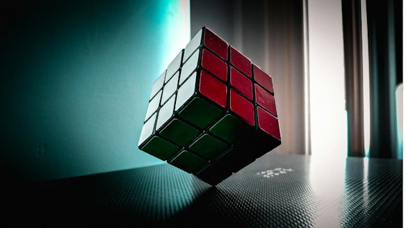 A Rubik's Cube is suspended in mid-air, captured in a softly lit room with light coming from tall windows. The cube displays red, white, and green stickers, and is positioned at an angle on a textured surface with a reflective sheen.
