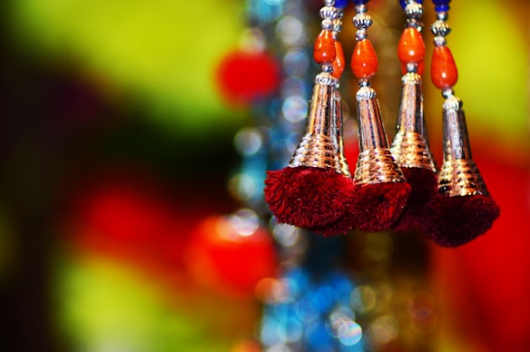 Four decorative tassels with intricate silver detailing and maroon fringes hang against a vibrant and colorful blurred background. The tassels also feature orange and silver beads above them.