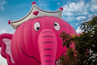 A large, pink hot air balloon shaped like an elephant with a cartoonish appearance, featuring big eyes and a smile. The balloon wears a white crown with red accents. The background includes a partly cloudy blue sky and the top of a tree with green and pink foliage.