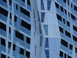 The image displays a modern architectural facade with angular and geometric patterns. Glass panels alternate with metallic sections, creating a textured and dynamic appearance. Reflective surfaces and varying shades of blue and gray contribute to the contemporary design.