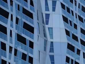 The image displays a modern architectural facade with angular and geometric patterns. Glass panels alternate with metallic sections, creating a textured and dynamic appearance. Reflective surfaces and varying shades of blue and gray contribute to the contemporary design.
