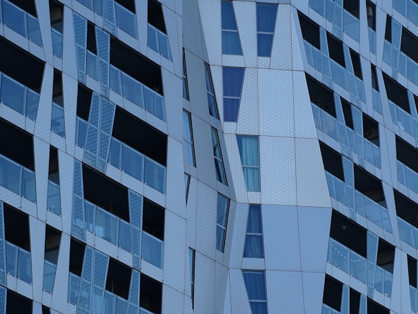 The image displays a modern architectural facade with angular and geometric patterns. Glass panels alternate with metallic sections, creating a textured and dynamic appearance. Reflective surfaces and varying shades of blue and gray contribute to the contemporary design.